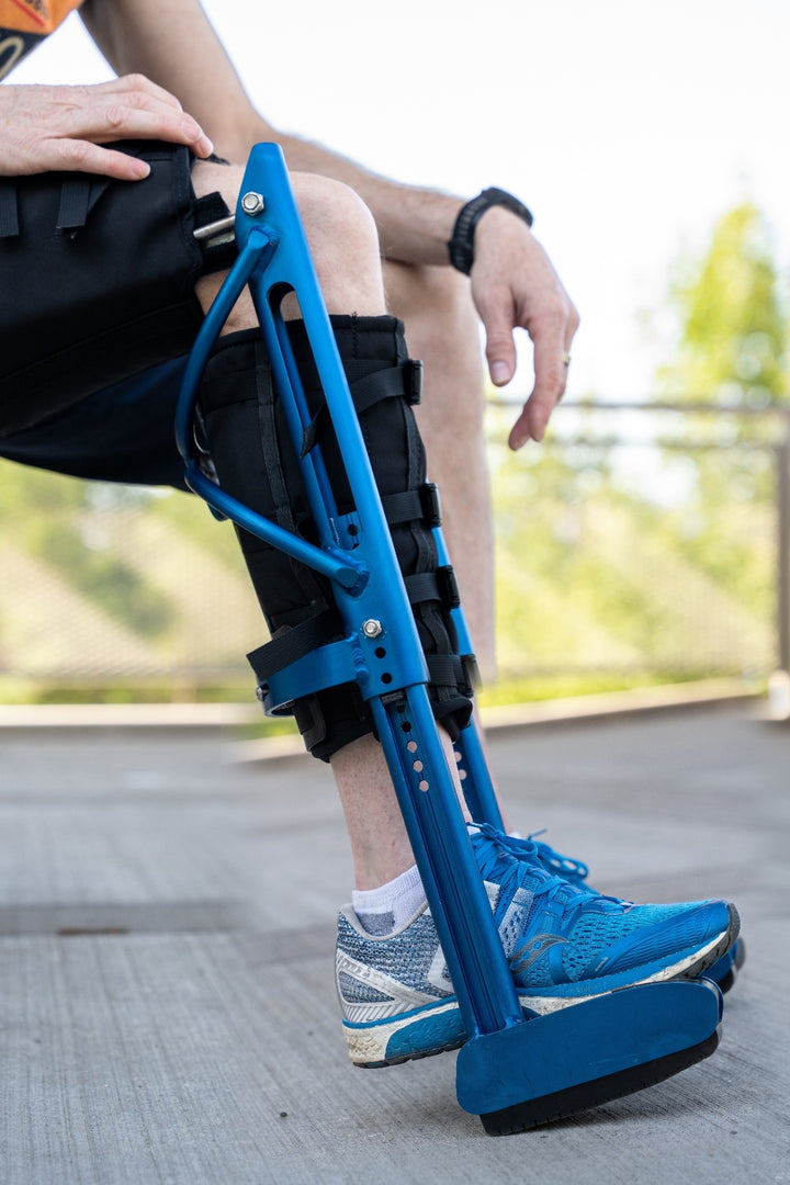 Close-up of a person sitting outdoors, wearing a blue XLeg hands free knee crutch on their right leg. The device is secured with adjustable straps, keeping the knee bent and foot suspended. This innovative knee crutch and crutch alternative provides stable, hands free mobility for lower leg injuries.