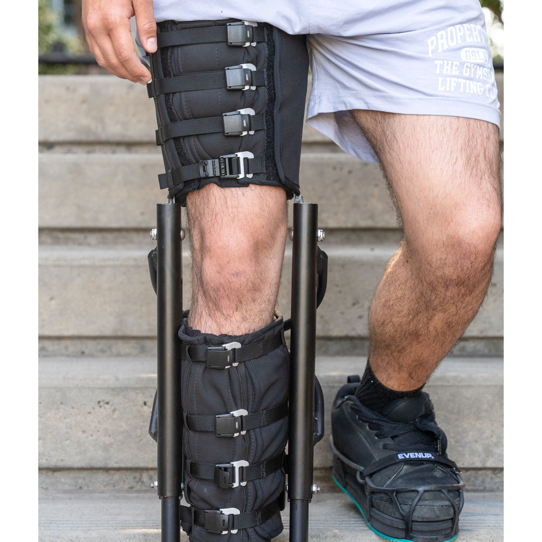 Close-up view of a person wearing the XLeg hands free knee crutch while standing on concrete outdoor stairs. The device features secure, adjustable buckled straps wrapping around the upper thigh cuff and lower leg support, keeping the knee comfortable and the foot suspended. The uninjured left foot is visible wearing a black sneaker equipped with a black level-up shoe attachment to balance leg height and maintain correct posture. This specialized knee crutch offers stable, hands-free mobility.