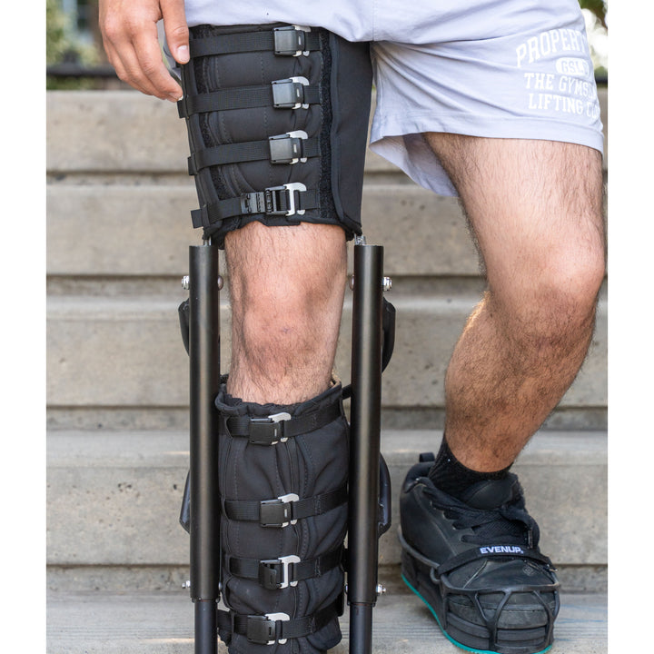 Close-up view of a person wearing the XLeg hands free knee crutch while standing on concrete outdoor stairs. The device features secure, adjustable buckled straps wrapping around the upper thigh cuff and lower leg support, keeping the knee comfortable and the foot suspended. The uninjured left foot is visible wearing a black sneaker equipped with a black level-up shoe attachment to balance leg height and maintain correct posture. This specialized knee crutch offers stable, hands-free mobility.