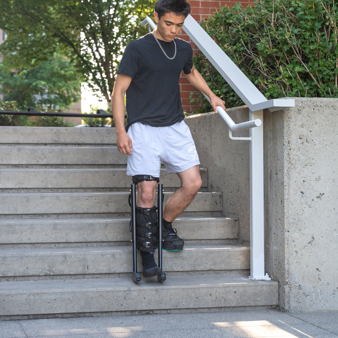 Young man descending outdoor concrete stairs while using a black XLeg hands free knee crutch. The device is securely strapped to his right leg, keeping the knee bent and foot suspended. His left foot wears a sneaker with an Evenup leveler attachment. This hands free crutch alternative provides safe, stable mobility for navigating steps.